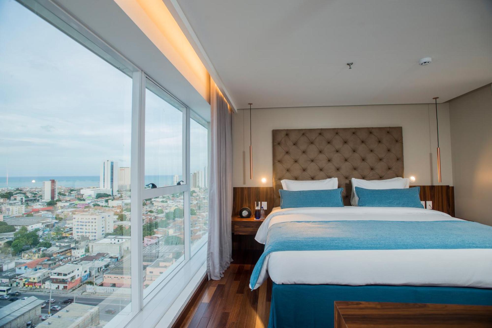 Hotel Luzeiros Ресифи Экстерьер фото The photo shows a modern hotel room featuring a large window with a city view. Inside, there are two neatly made beds with light blue and white bedding, flanked by wooden nightstands. The room has a contemporary design, with a padded headboard and so